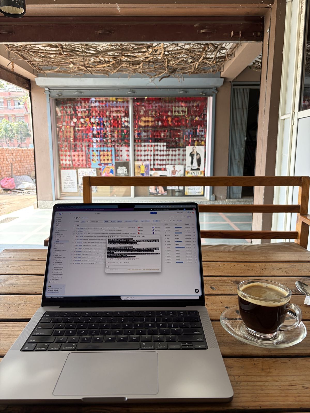 A laptop showing a bug tracker board beside a glass cup of black coffee on a wooden café table