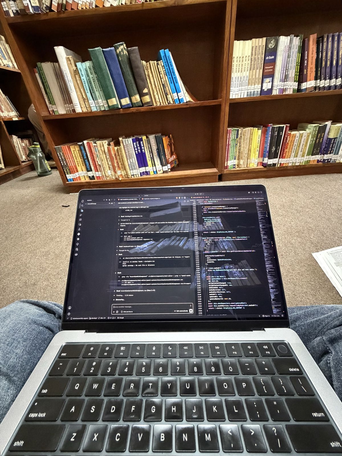 A laptop balanced on knees in front of wooden library shelves full of books