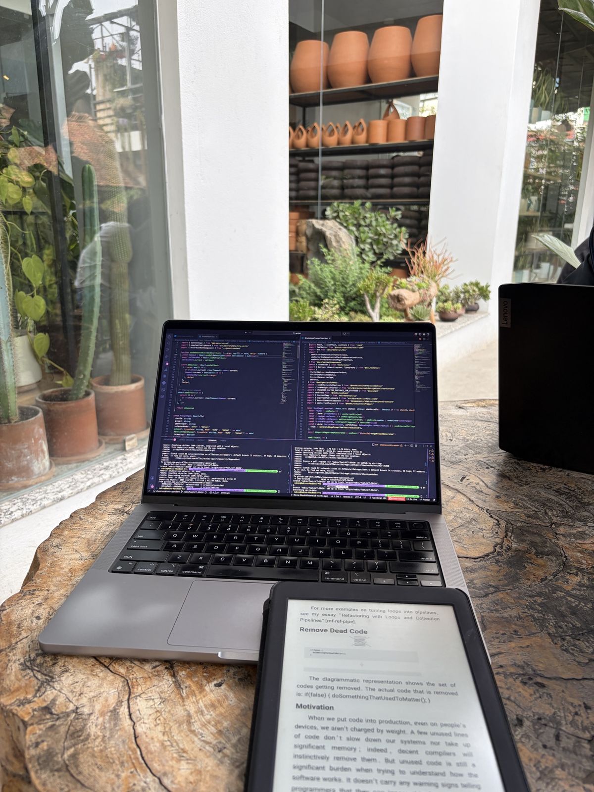 A laptop and an e-reader showing the Refactoring book on a live-edge table, surrounded by potted plants and terracotta pots