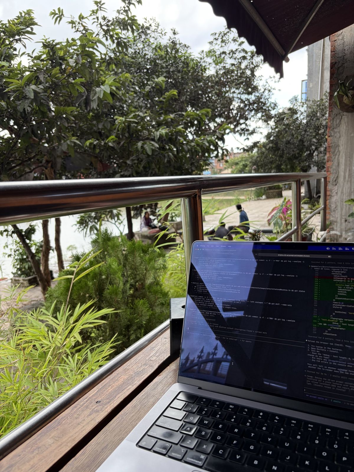 A laptop open on a balcony rail, looking out over greenery and a pond
