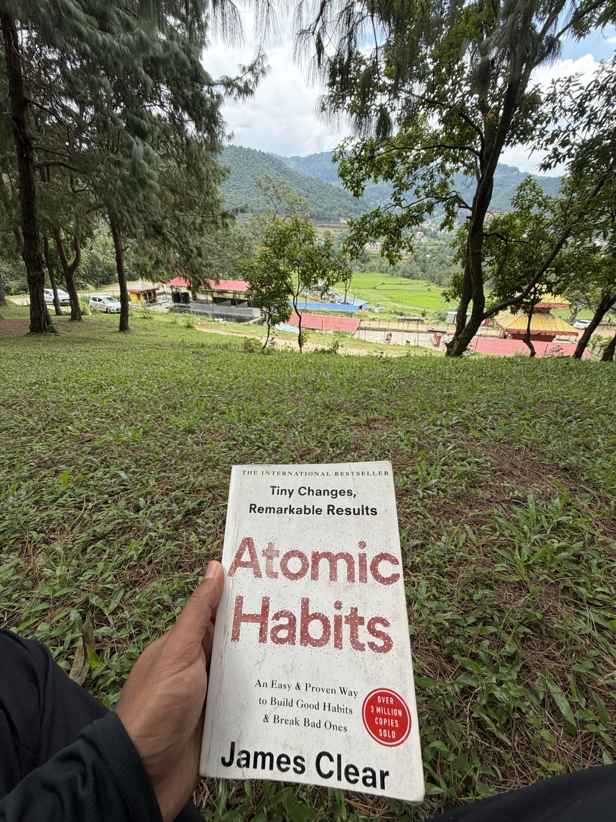 Holding a copy of Atomic Habits on a grassy hillside looking over a green valley